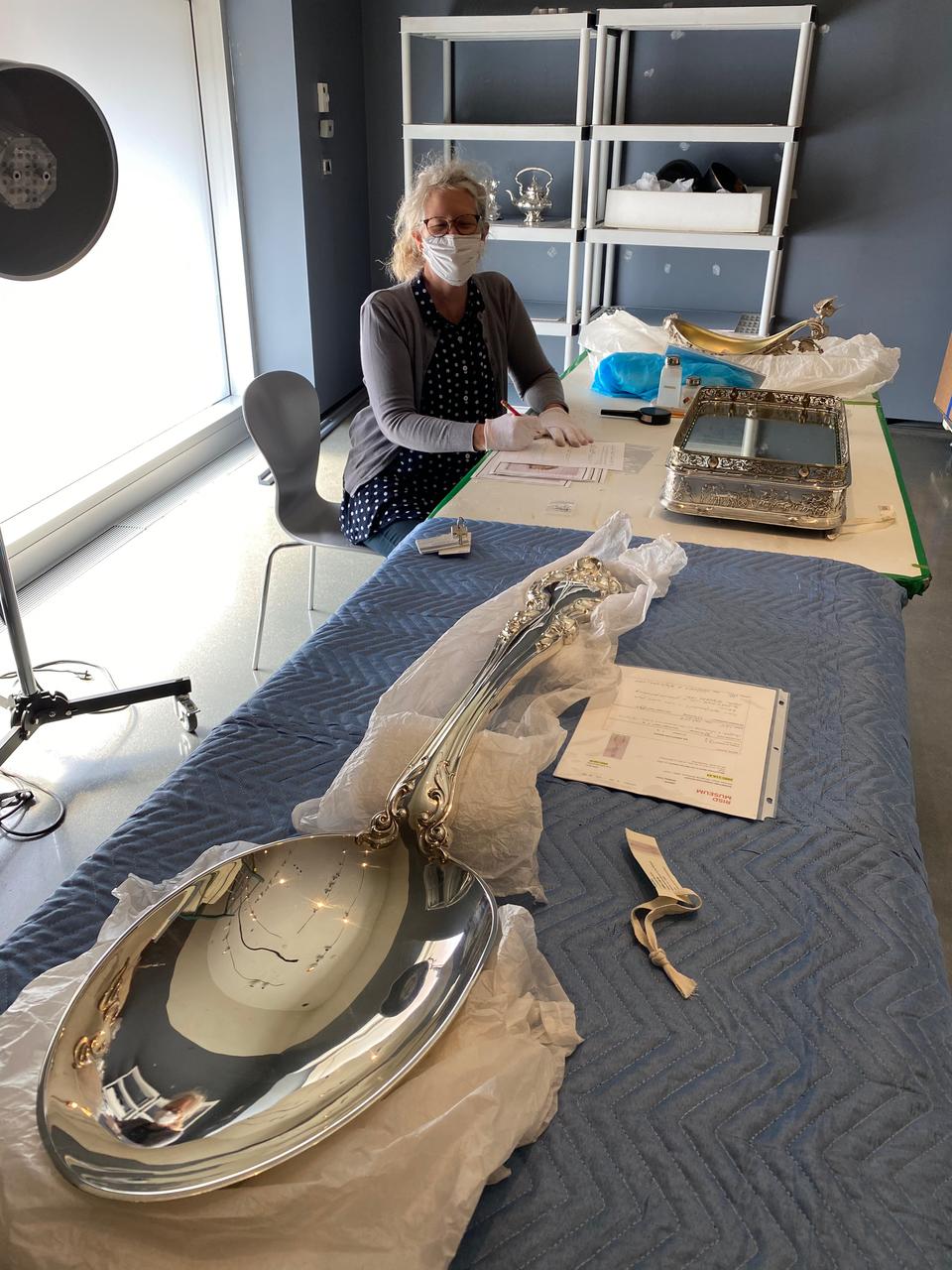 A masked museum worker sits at a table with several objects. The work dominating the foreground is an enormous shiny silver spoon.