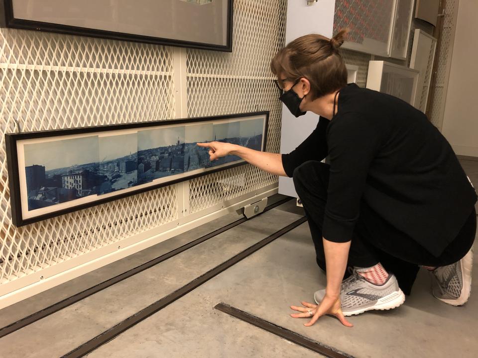 A masked young person crouches to get a closer look at a long horizontal work hanging at the bottom of a rack. They point at a detail in the image.