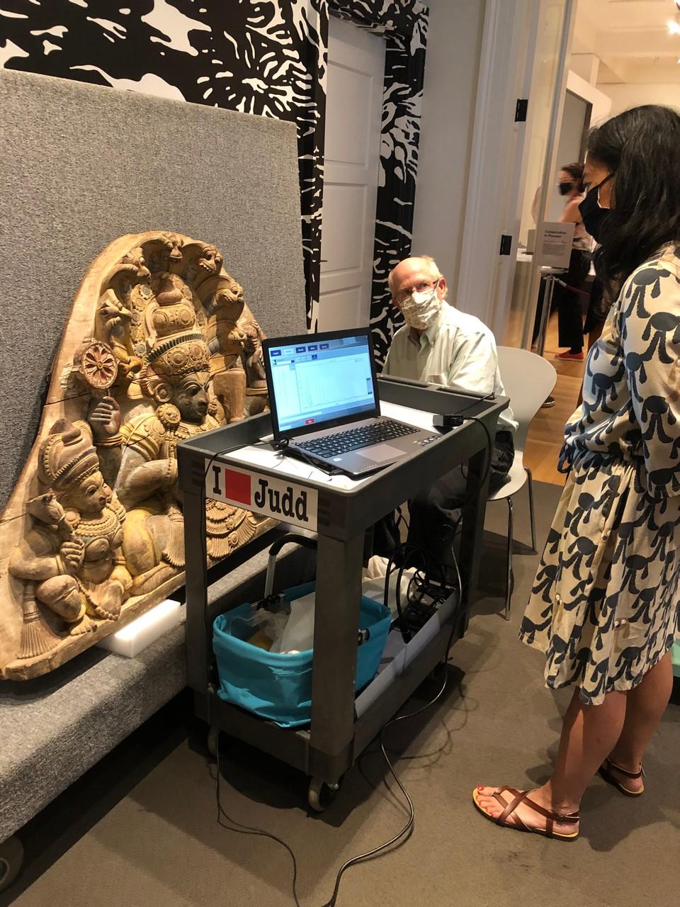 A masked person sits beside a carved stone artwork and a computer on a cart. They interact with a standing person, also masked.
