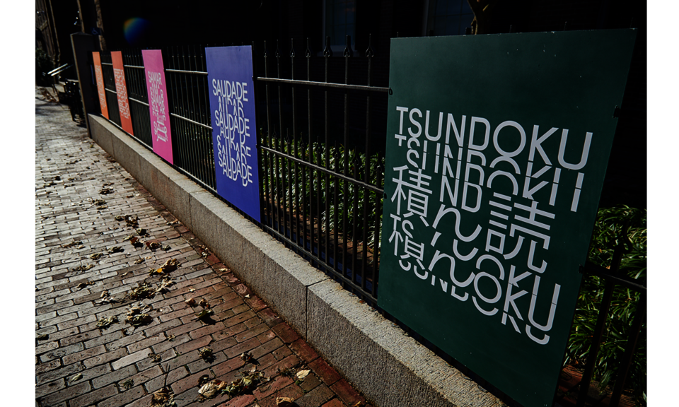 Beside a brick sidewalk, several signs are attached to a wrought-iron railing. The first sign, in green, reads “Tsundoku,” followed by stylized text.