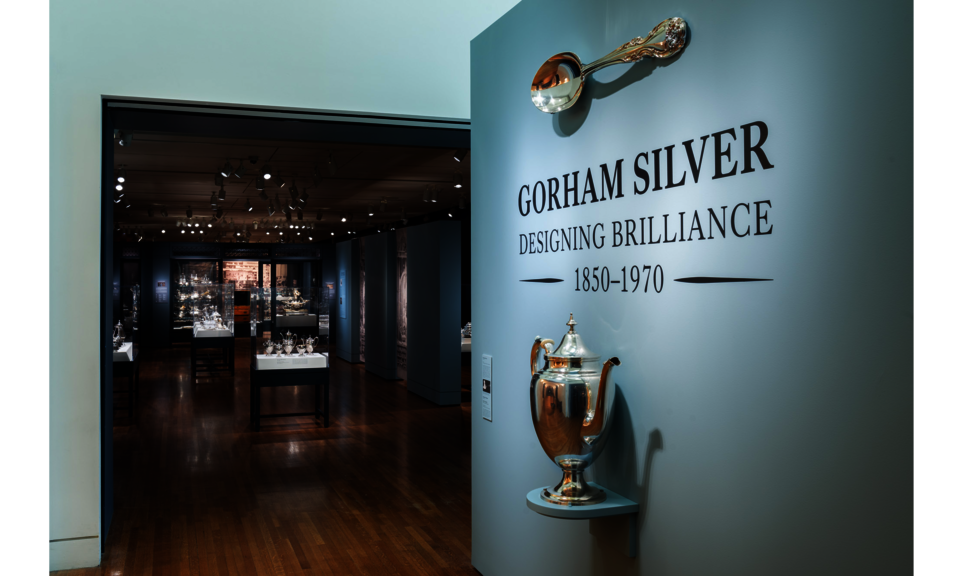 Text on a light gray wall just outside an exhibition space says: “Gorham Silver: Designing Brilliance 1850-1970.” Mounted on the wall are an oversized silver coffeepot and spoon.