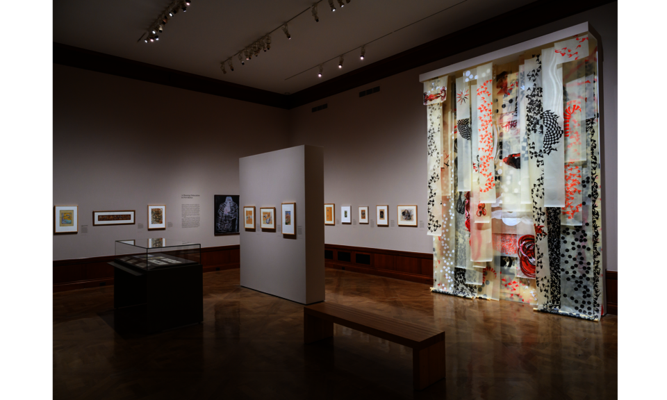 The walls of a large gallery space are hung with many works of art. At right is a colorful floor-to-ceiling installation of various decorative panels.