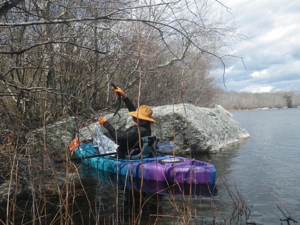A person seated in a kayak on calm water. The trees above them and the vegetation we view them through are leafless. The sky is cloudy.