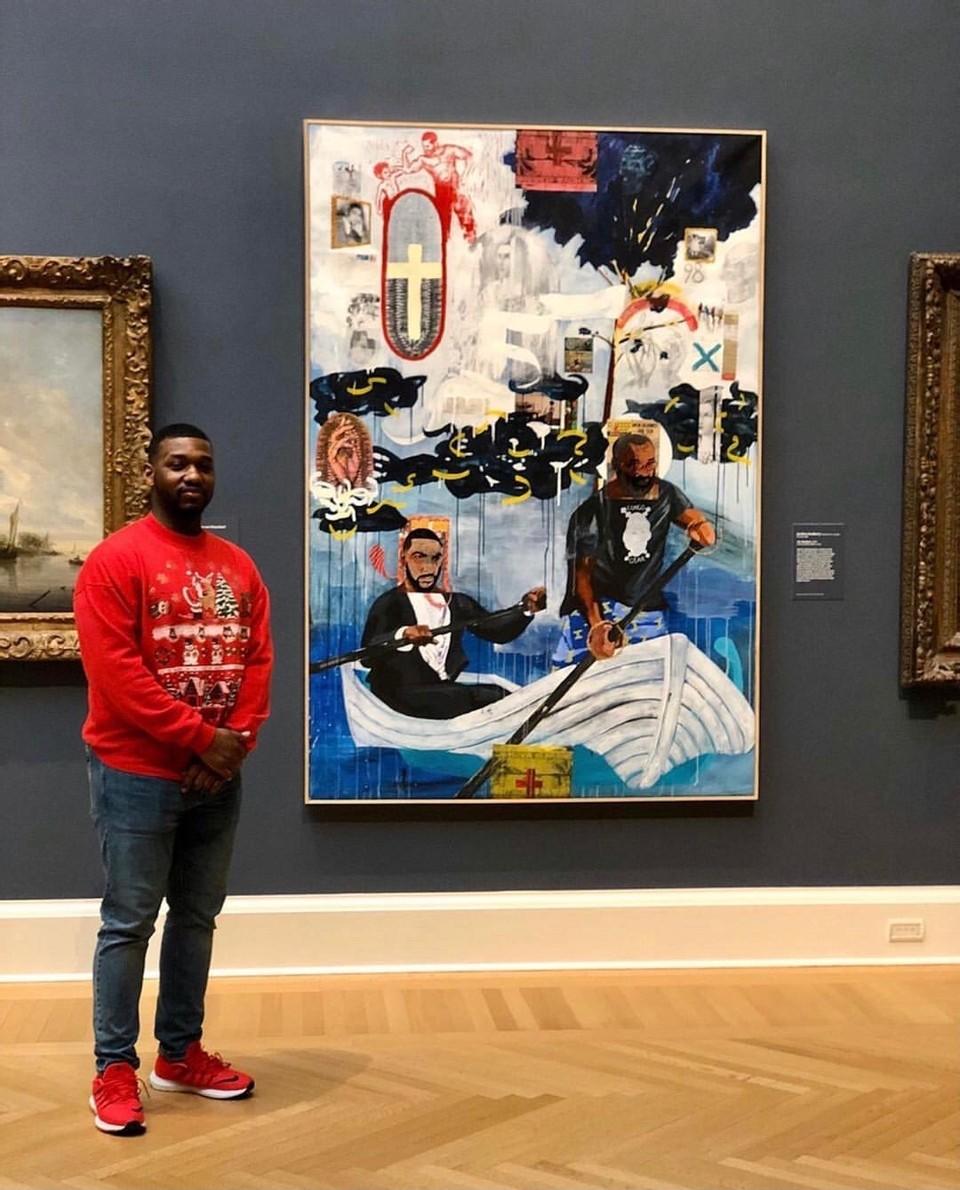 A young dark-skinned person stands smiling in a gallery. They are beside a large, expressive painting depicting, among other things, two dark-skinned people rowing a boat.