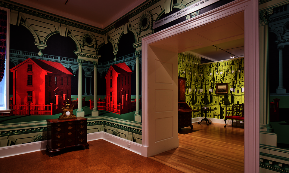 Colorful wallpaper embellishes the walls of two adjoining rooms containing antique furniture. The foreground pattern includes large vibrant red houses and green architectural details. A chartreuse pattern is adjacent.
