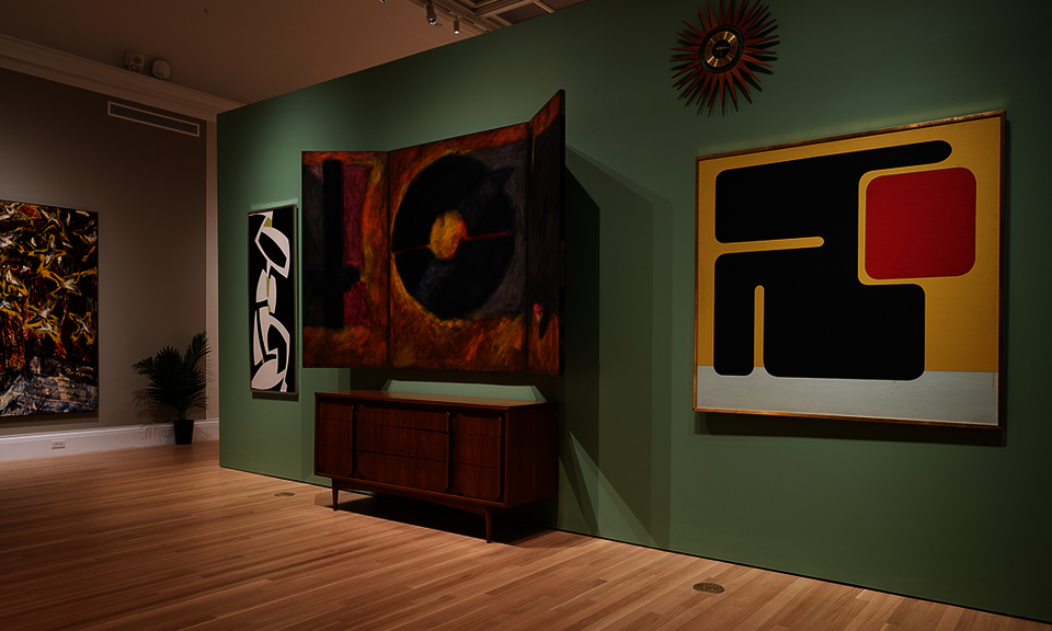 Modern paintings and furniture are displayed in a gallery space with pale green walls and wooden floors.