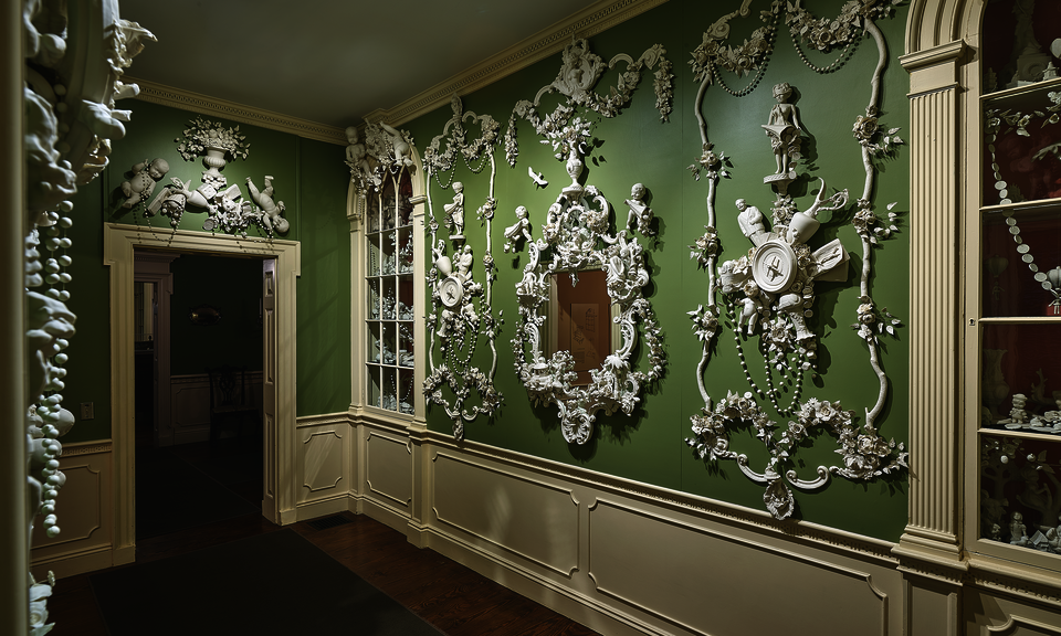 The green walls of a stately interior space featuring cream-colored woodwork are elaborately decorated with mirrors and delicate white three-dimensional figural embellishments. A built-in glass-doored cabinet is at right. 
