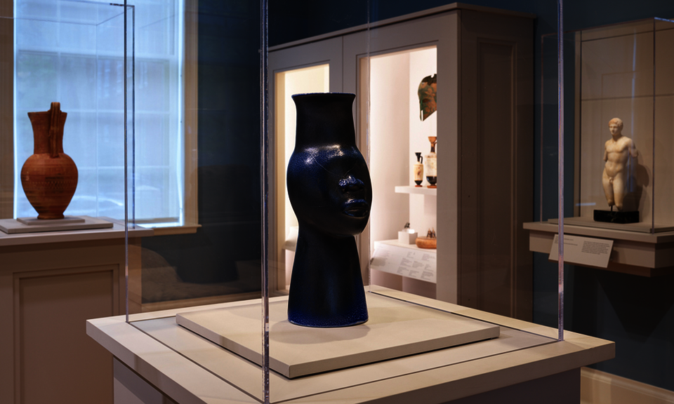 In a case in a gallery’s center is a sleek black vessel shaped like a stylized head with an elongated neck. Cases along the perimeter contain other objects.