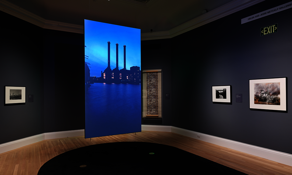 Hanging in the center of a gallery with dark-gray walls is a screen. The projected image shows an industrial building with three smokestacks. Artwork hangs on the surrounding walls.