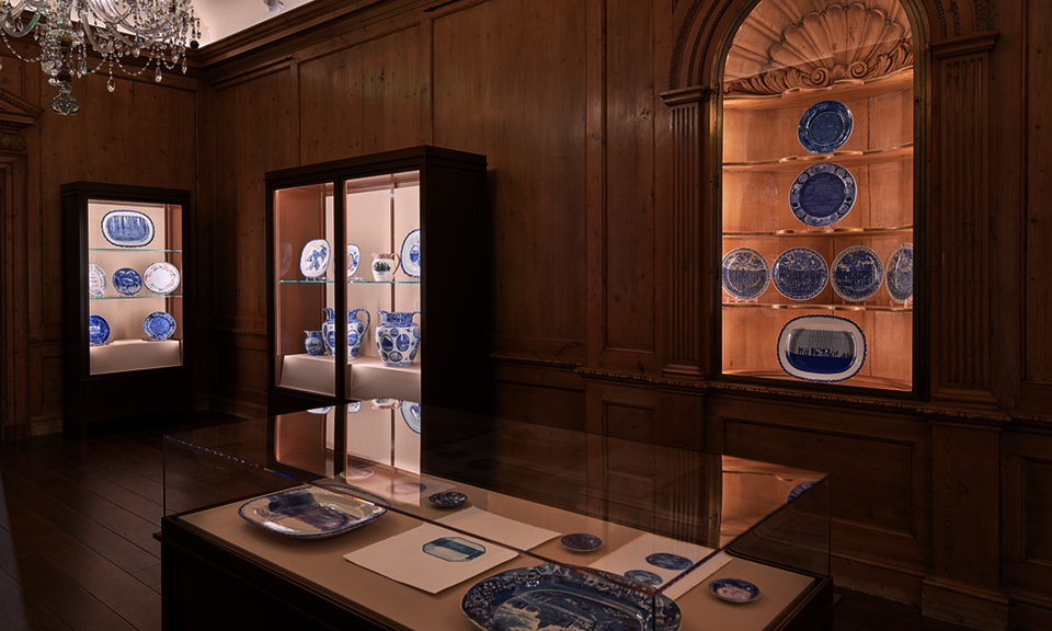 Blue and white ceramics are displayed in cases along the walls of a wood-paneled gallery. A case in the foreground contains additional works. A crystal chandelier hangs at top left.