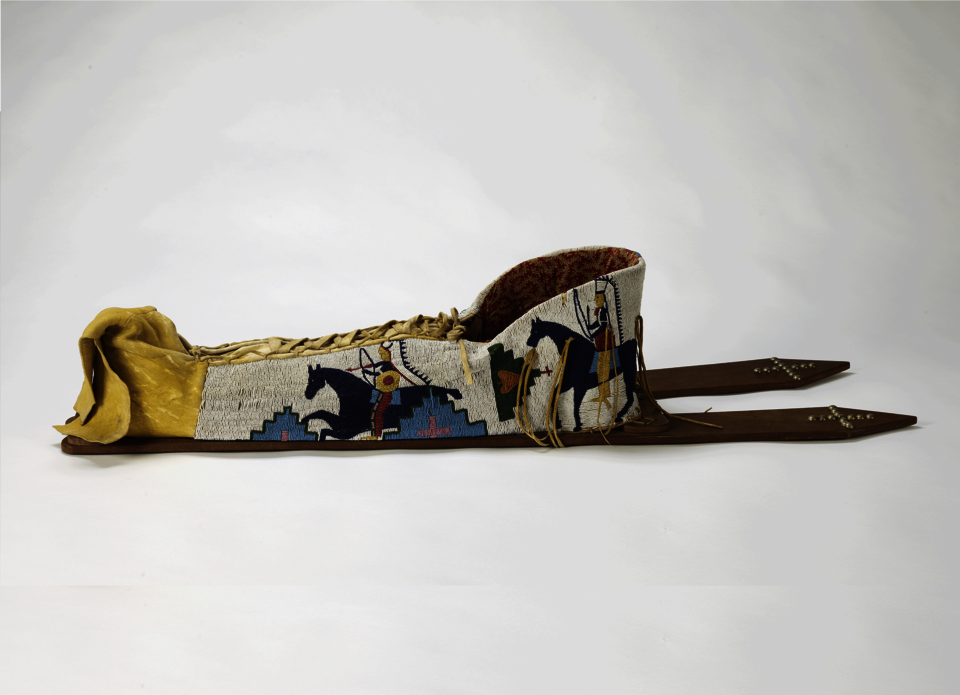 Beaded baby carrier lying flat on a surface. Back boards extend to the right. Beadwork designs in blue on white background depict horses, riders, and geometric decoration.