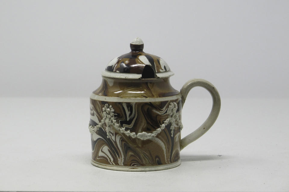 An earthenware mustard pot that is white, light brown and dark brown. The lid and handle are rounded and there are swag floral decorations on the body.