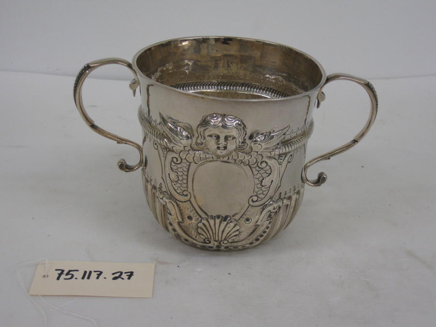 A silver cup with symmetrical handles, swirling raised designs and a small raised face on the upper half of the cup.
