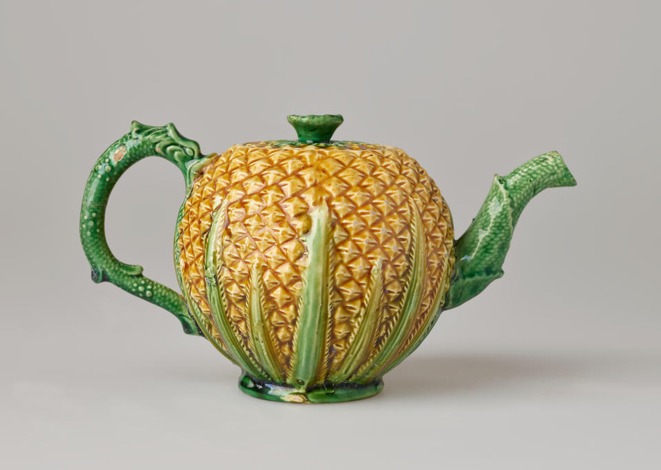 A sculptural teapot with a yellow body and texture similar to a pineapple, green handle, foot, lid, and spout with a floral leaf or stem like decoration and texture.