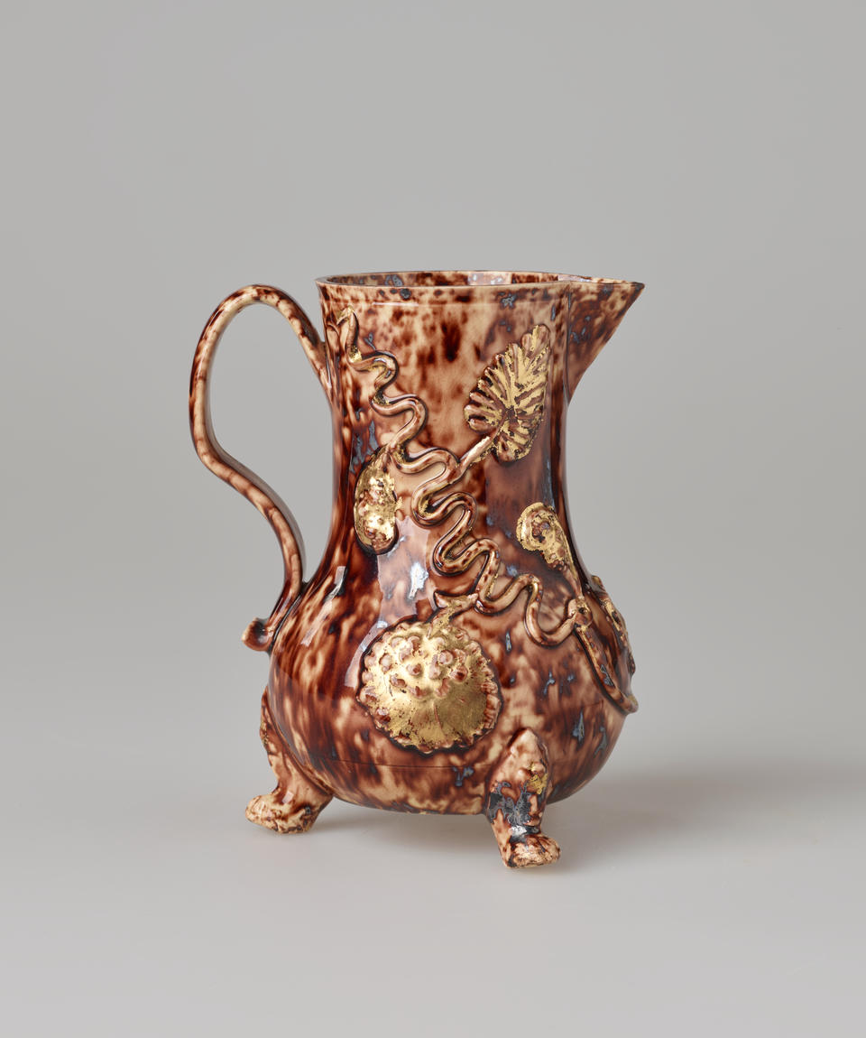 A brown ceramic pitcher with a spout, handle, and multiple delicate feet. There are swirly floral decorations.