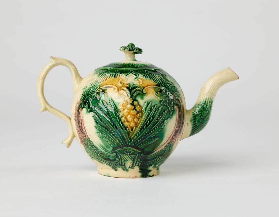 A bulbous ceramic teapot with a long curved spout and handle, white, dark green. and yellow glaze, and floral decorations ranging from leaves to a more central pineapple.