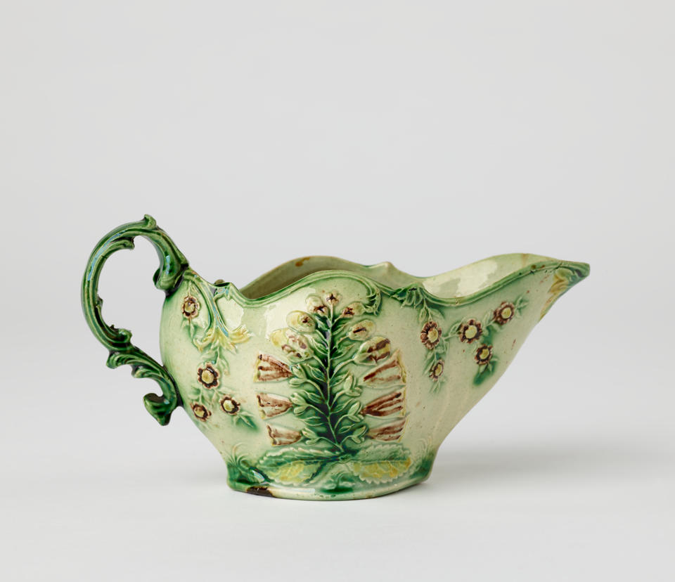 A short boat shaped pitcher green, cream, and brown in color with a sculptural handle and floral decorations.