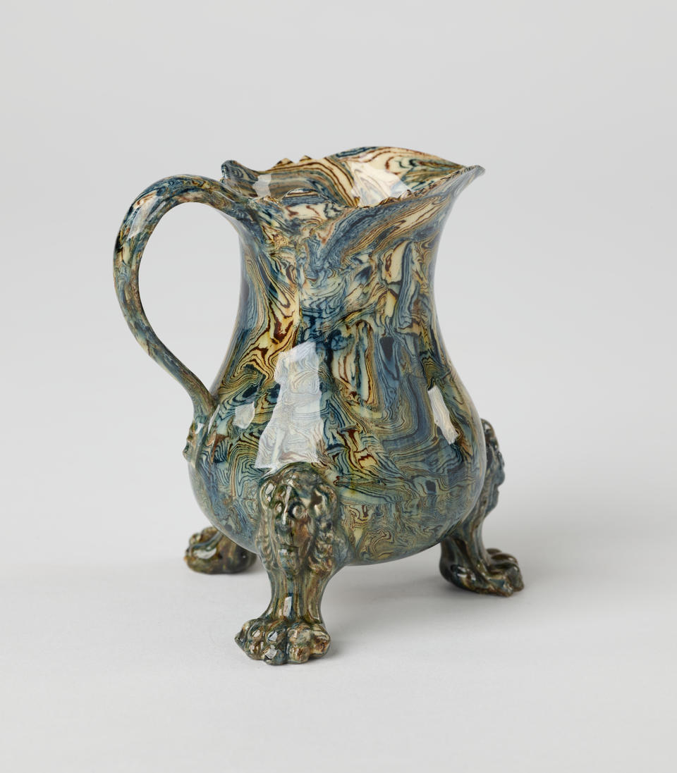 An agate wear vessel with a handle, three sculptural feet, and a spout. The vessel is shiny and has cream, blue, and green swirling colors.