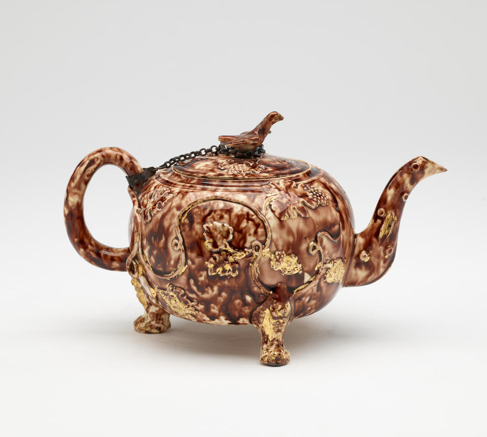A dark brown and white ceramic teapot with protruding feet, a spout, handle, and lid. The finial on the lid is connected to the handle by a chain.