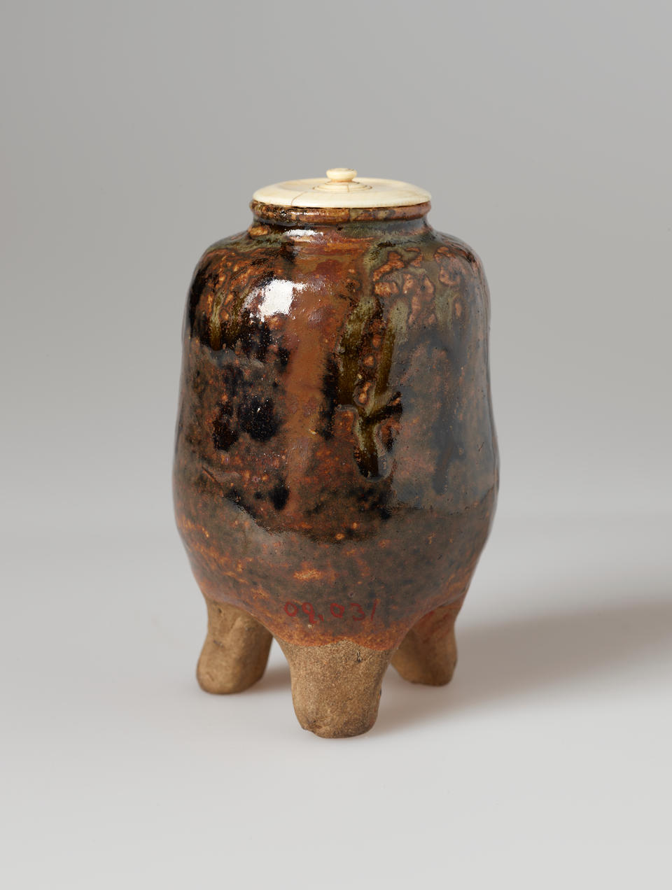 Small vessel in shades of reddish brown and black, standing on three rounded feet, with a cream colored lid.