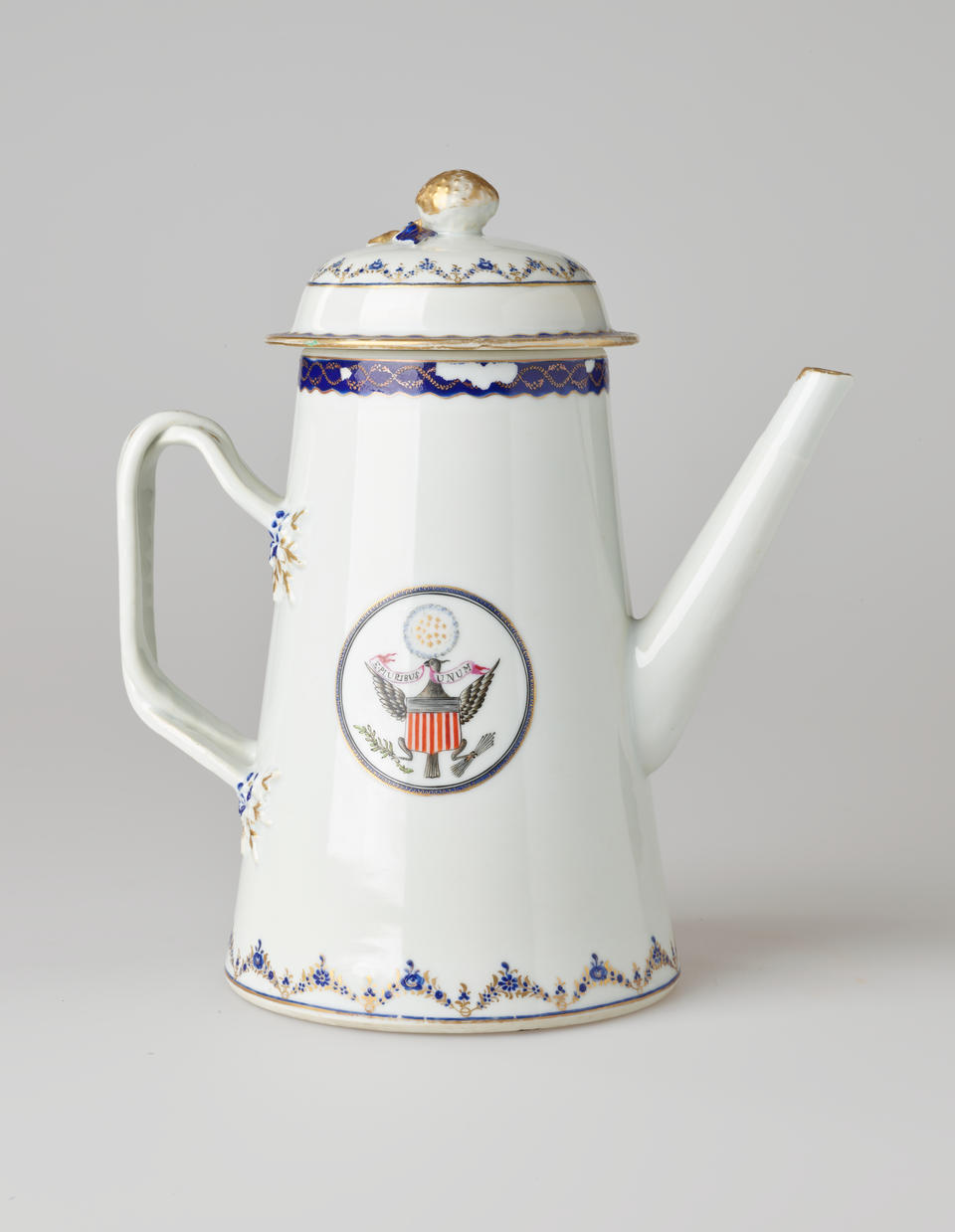 A white ceramic coffee pot with dark blue and gilded decorations, a straight spout, angular handle, and heraldry imagery in a circle on the body of the coffee pot.