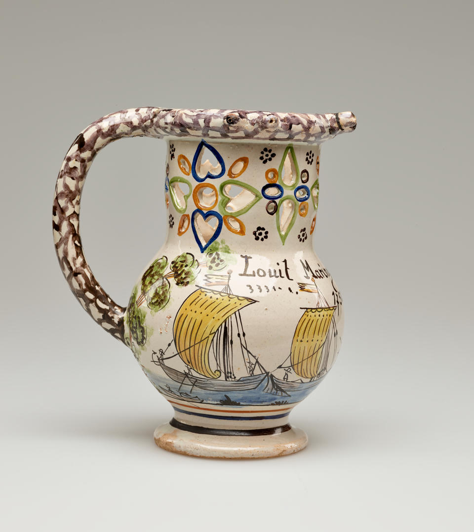 A jug with a rounded handle and pierced designs. The cream colored body is decorated with images of ships and a tree. 