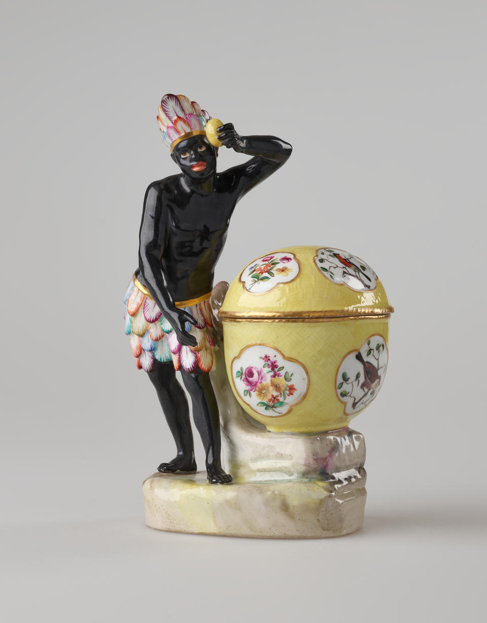 A sculptural figure with glazed black skin, a colorful headdress and bottoms. This figure is standing next to a bright yellow covered sugar basket with floral decorations. 