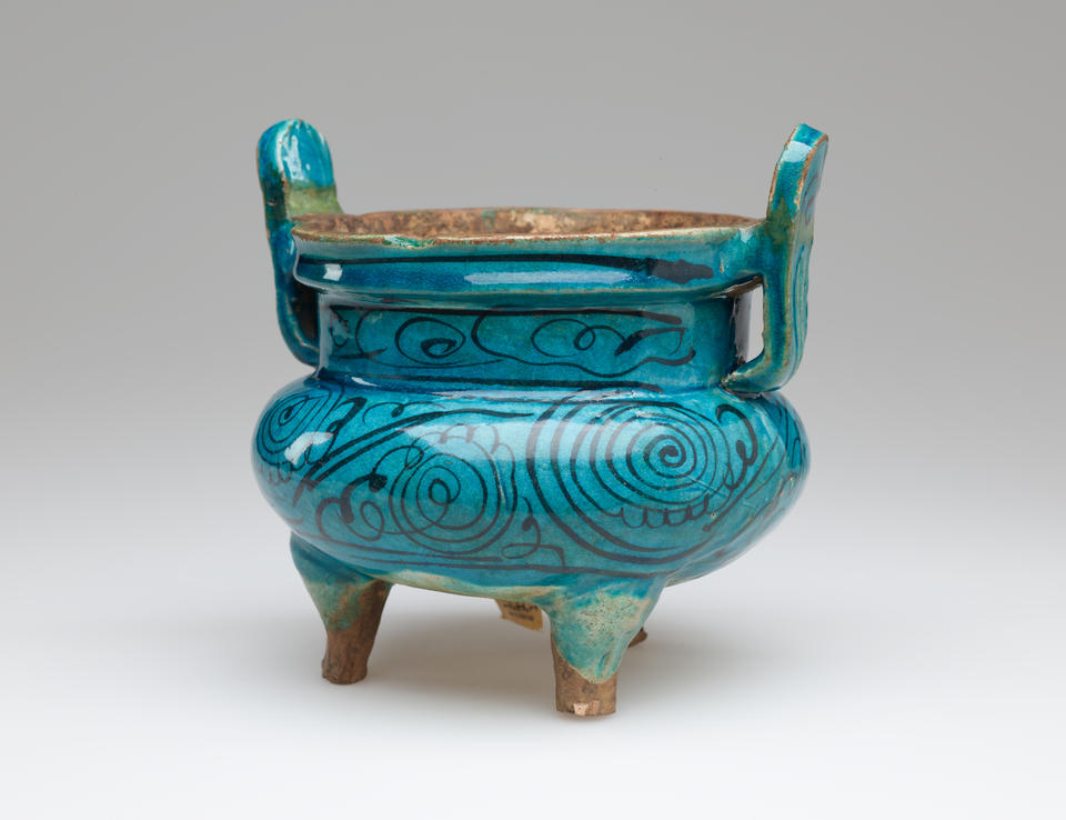 BP- Round, blue vessel with dark thin detailing on body. Two thin handles flare upward, and has four unglazed feet. 