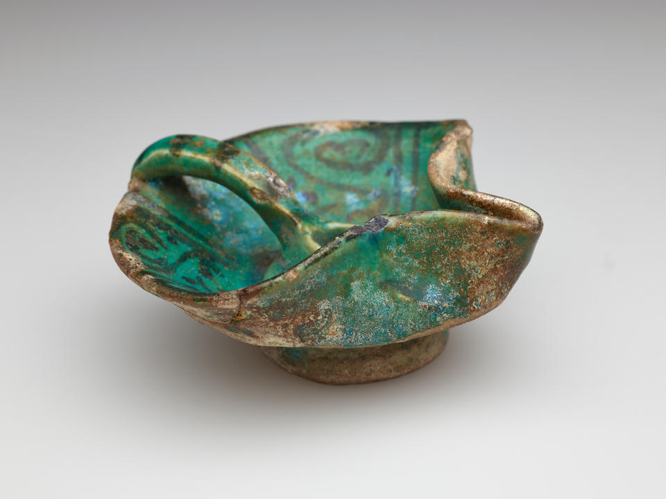 An irregularly shaped blue-green iridized vessel with dark designs. Rounded on one side with a handle that comes to the center of the vessel. The other side has a folded spout-like edge.