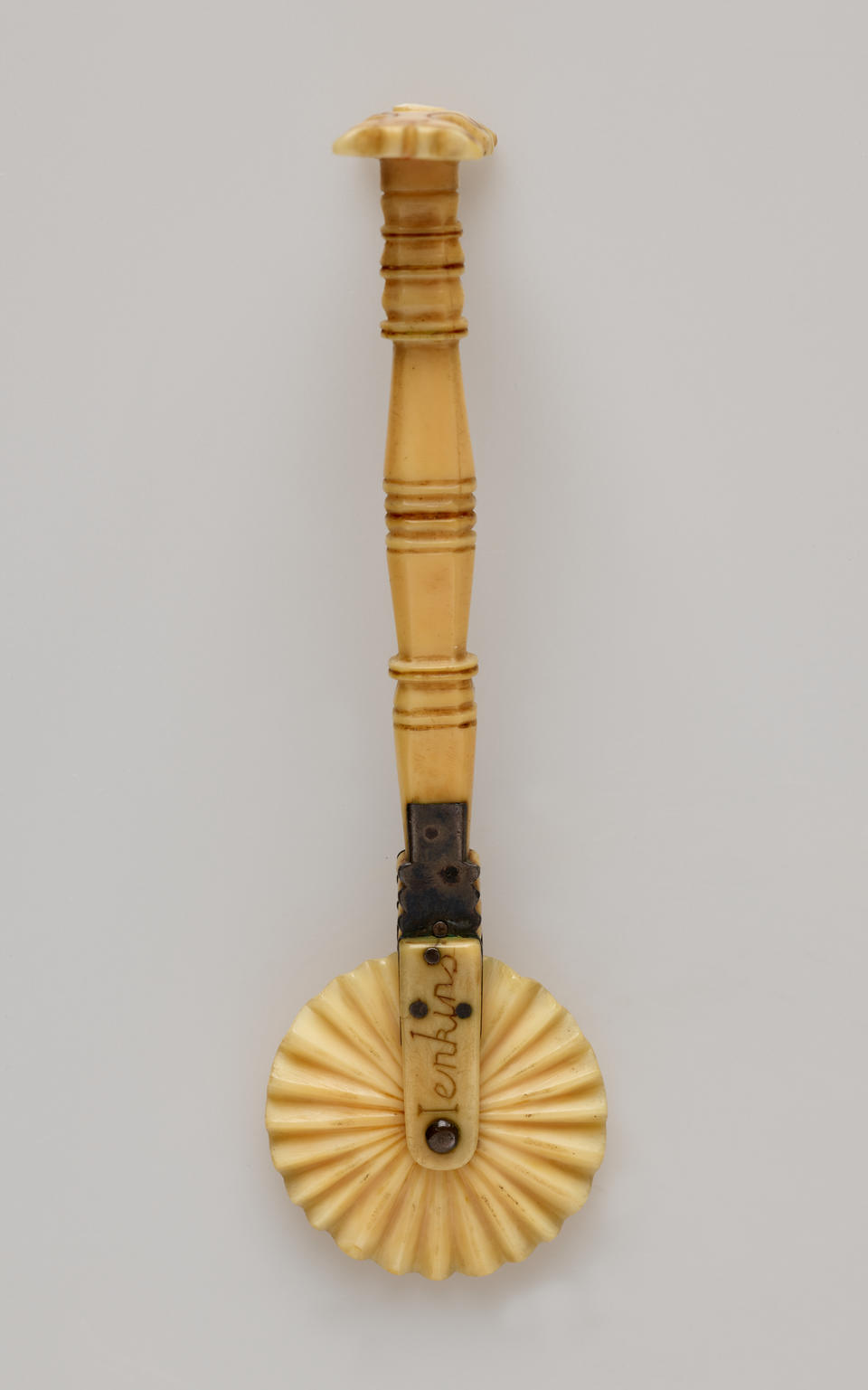An ivory pie crimper with a decorative handle and a textured wheel. There are letters on the part attaching the wheel to the handle.