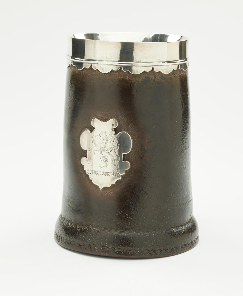 BP: A leather, silver, and pewter tankard. The body is mostly dark with the exception of a decorative crest, rim and interior. 