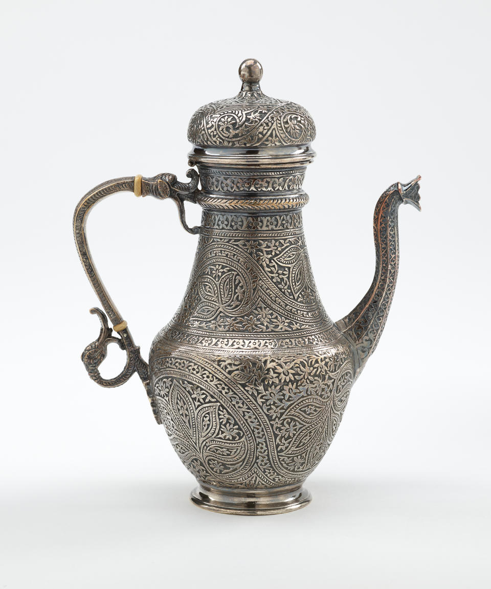 A silver and ivory coffee pot with detailed delicate decorations, handle with a lid hinged to it, and spout.