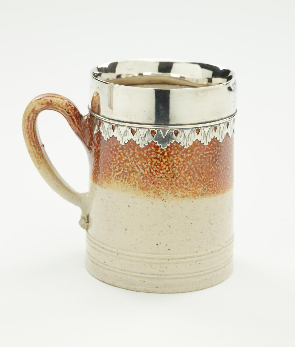 A salt glaze and silver mug with vertical decorative elements and one handle.