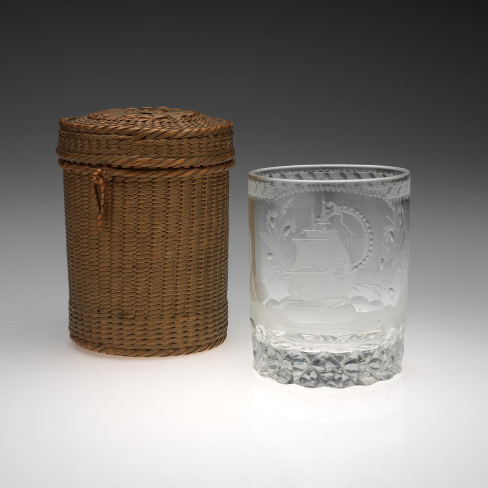 Rounded clear glass with etched designs next to a brown woven basket of the same shape. A small, tan-colored implement lays to the left of the basket and glass. 