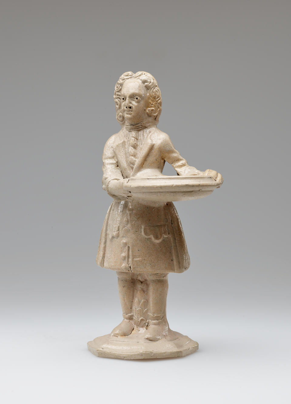 A sculptural figure standing, holding a vessel, dressed in historical clothing with tightly curled hair. The entire stoneware figure is a light cream color.
