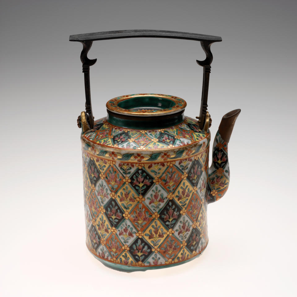 A cylindrical teapot with an elaborate design of flowers inside diamond shapes of varying colors. A thin, square shaped handle with a flat top sits above the teapot.