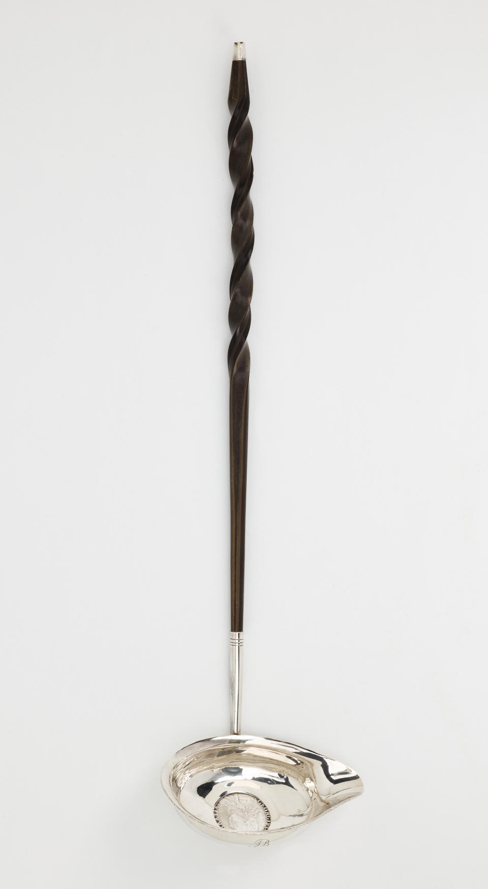 A silver and baleen coin ladle. The handle is twisted and dark and the ladle is silver with a small coin in the center. 
