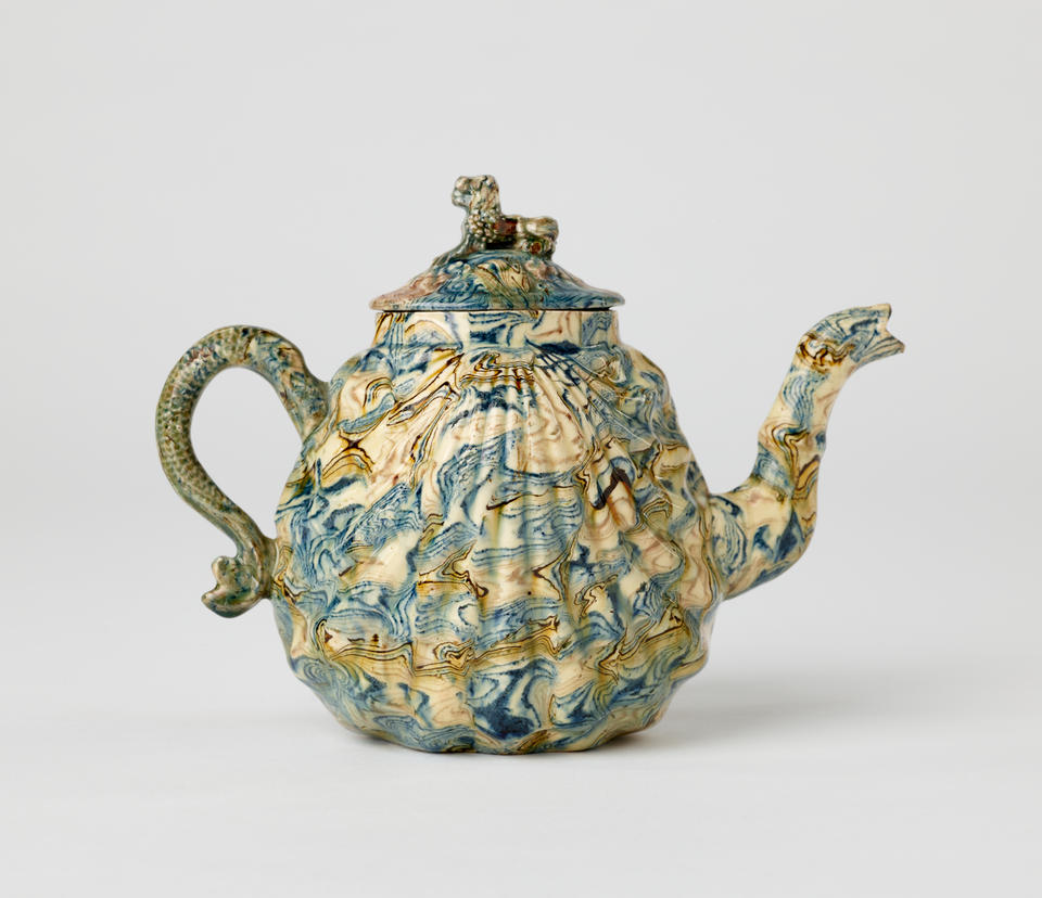 A sculptural agateware teapot, with a sculptural fin handle, shell shaped body, decorative spout, and animal sculpted finial for the lid. It is blue, brown, and cream in color.