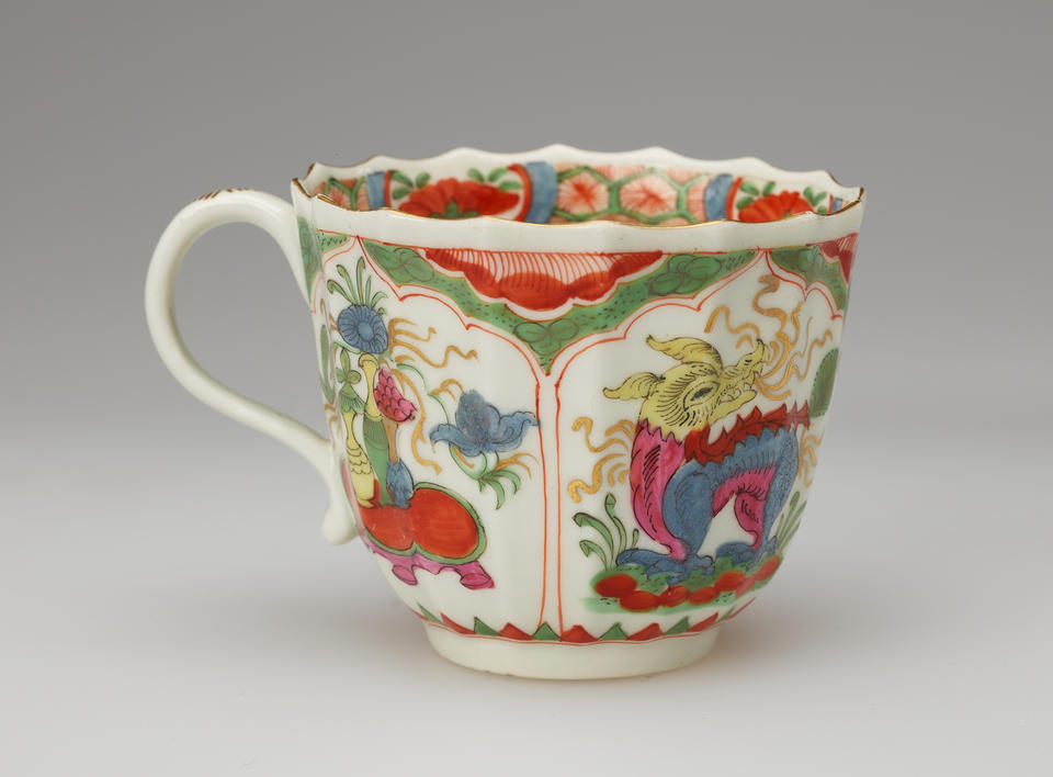 A white cup with a scalloped lip, green, red, blue, pink, yellow, gilded decorations, and two vignettes: an animal and the other a table with vessels on top of it.