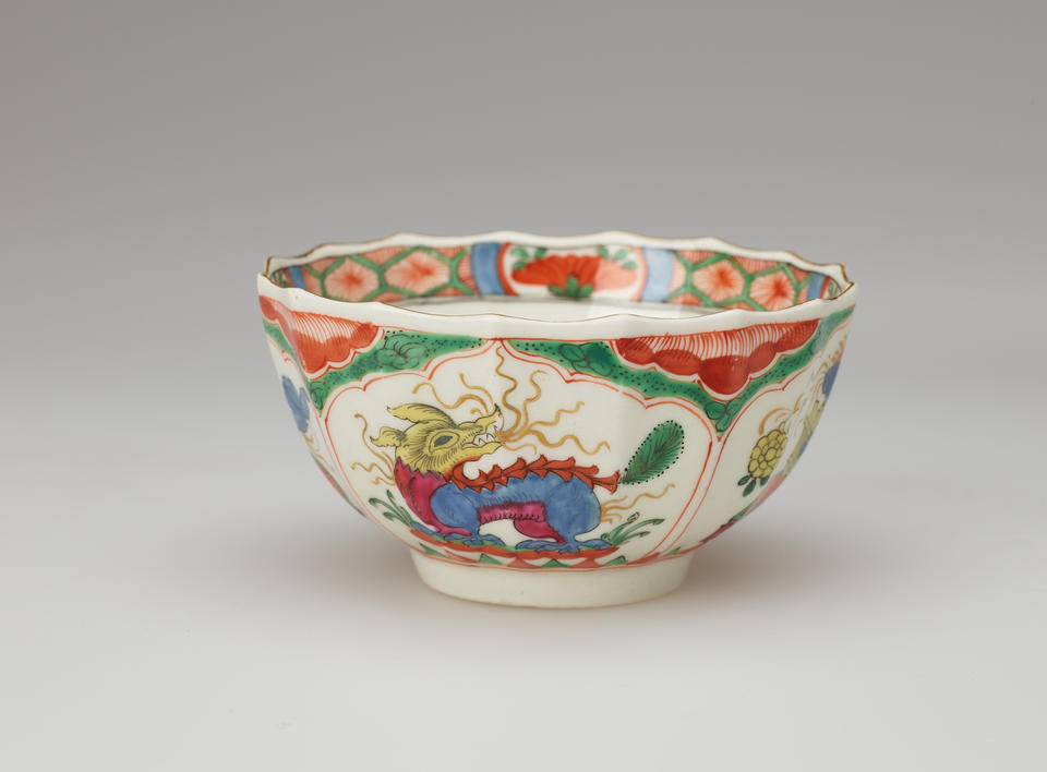 A white tea bowl with green, red, blue, pink, yellow and gilded decorations. The lip of the vessel is scalloped. 