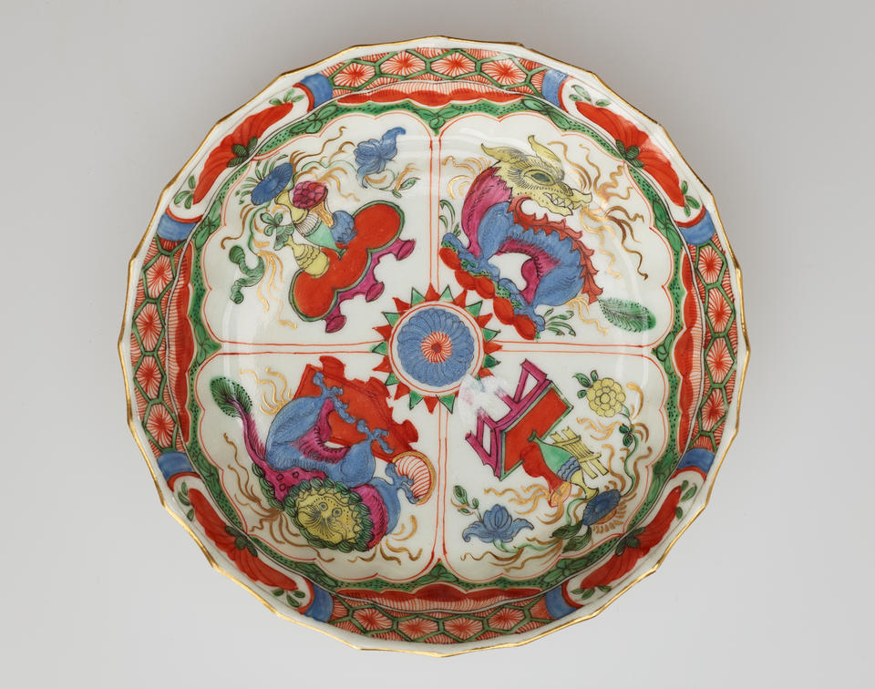 A white saucer with green, red, blue, pink, yellow, and gilded decorations. Decorations separated into four vignettes; two animals and two scenes of a table and vessels containing floral elements.