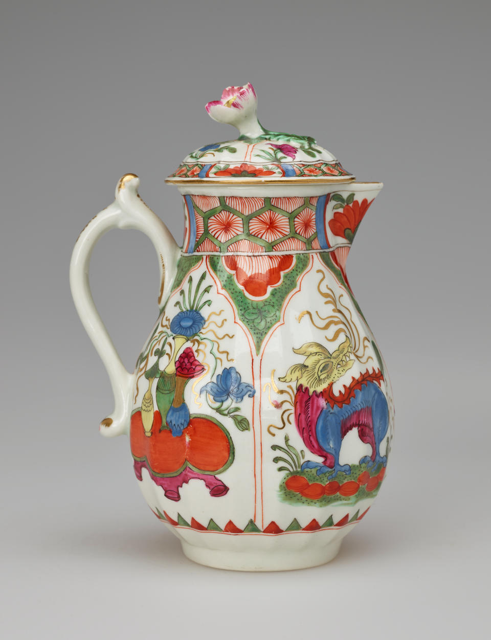 A white milk jug with a handle, lid, and spout with green, red, blue, pink, yellow, gilded decorations,and two vignettes; an animal and a table, vessels, and floral elements.