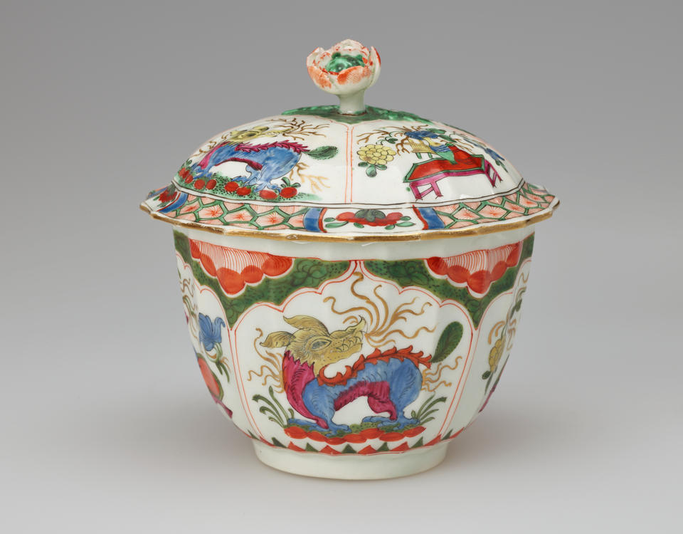A white sugar bowl with lid and finial with green, red, blue, pink, yellow and gilded decorations. The finial is a sculptural flower white, red and green.