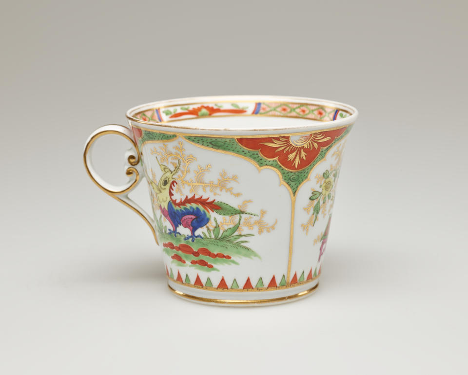 A chocolate cup that is with green, red, blue, pink, yellow, gilded decorations, and vignette with a dragon on a table surrounded by gilding.