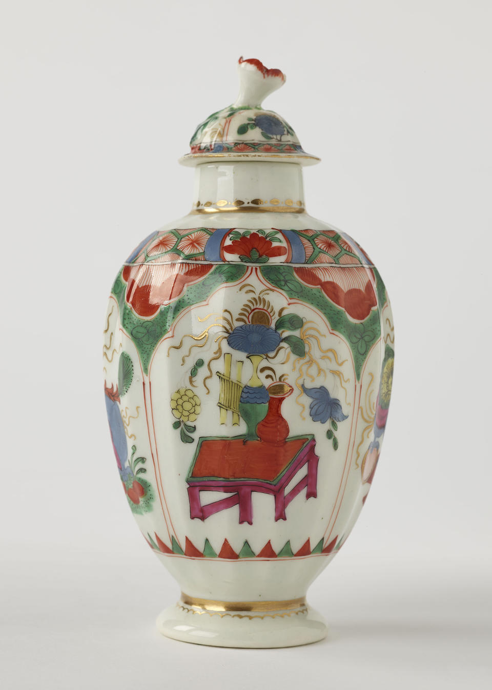 A porcelain tea caddy with a lid and green, red, blue, pink, yellow and gilded decorations. The finial is a sculptural flower white, red and green