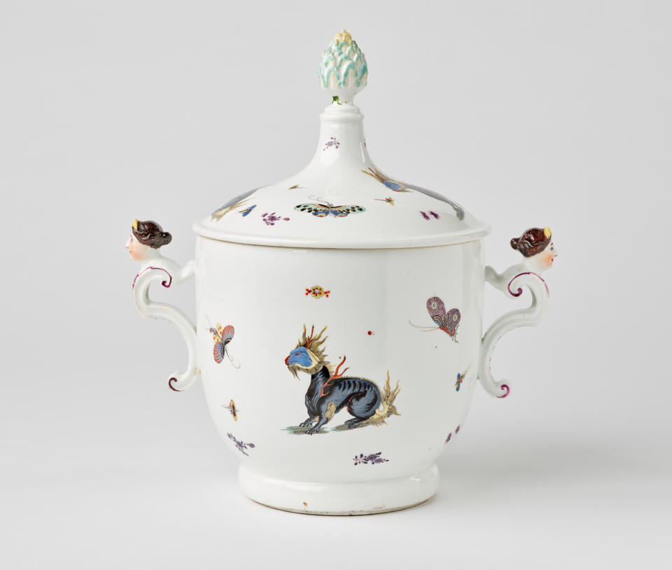 A white porcelain icecream pail with dragons and bugs decorations, there are two sculptural handles that are figure’s heads, the lid has a sculptural finial.