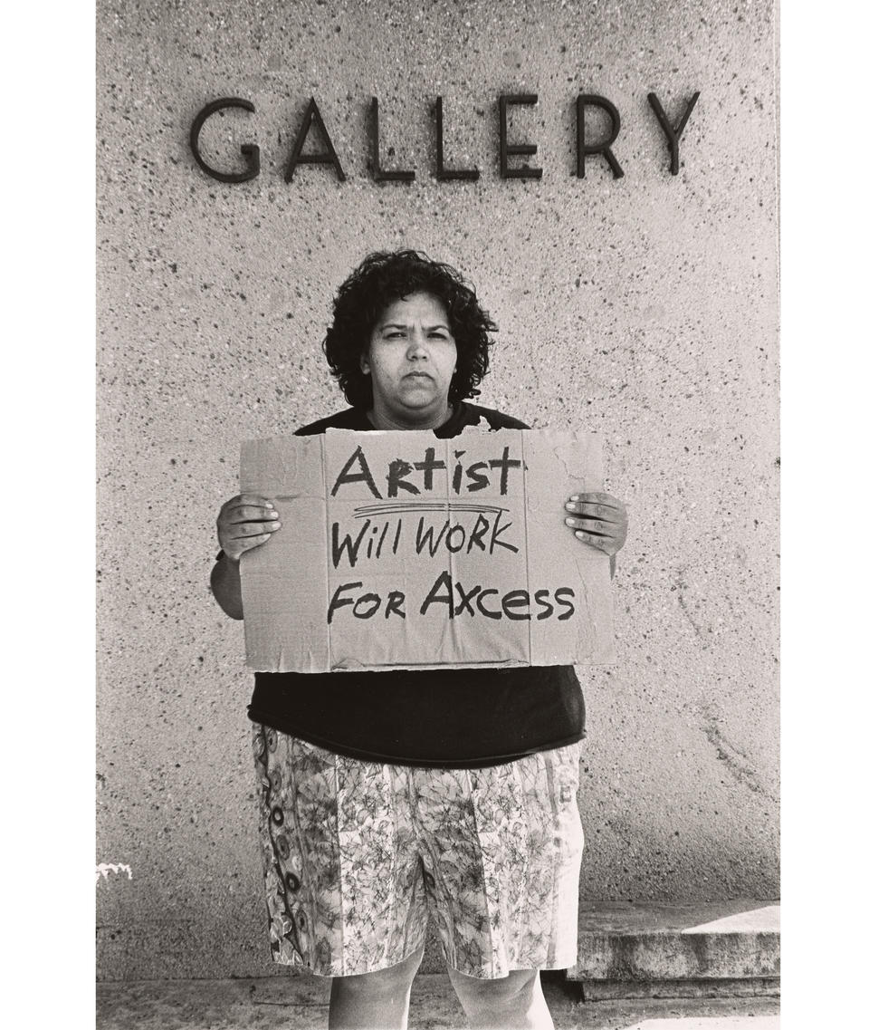Black and white photo of an unsmiling woman looking at the camera and holding a sign that says “Artist. Will work for axcess.” Lettering on the wall above says “Gallery.”