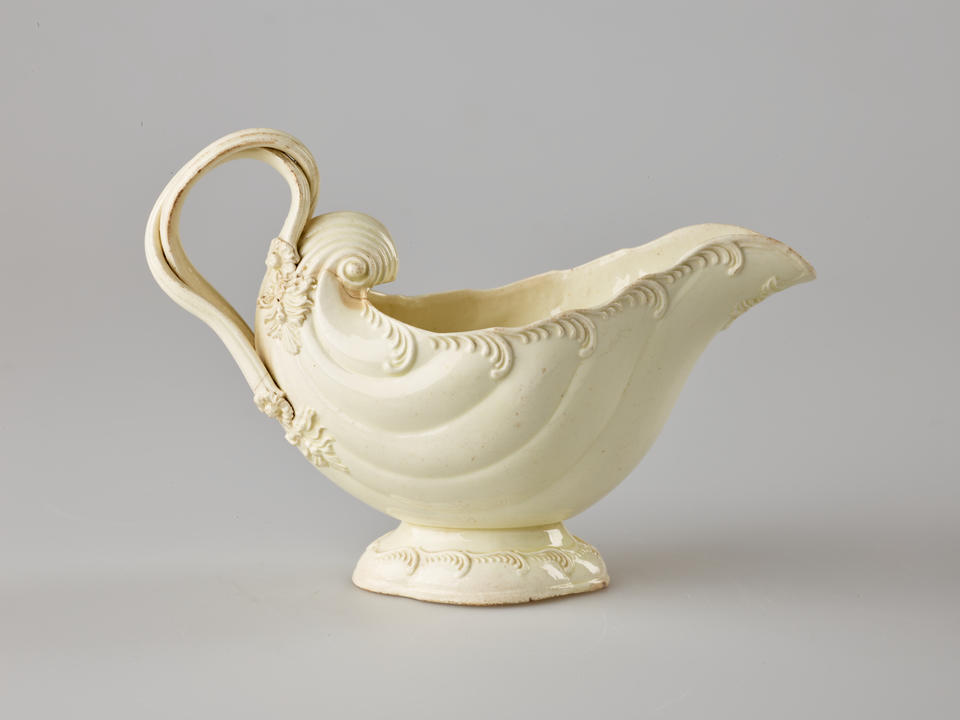 A cream colored vessel with rounded foot and handle. The body of the vessel is shell shaped and has swirled decorations. 