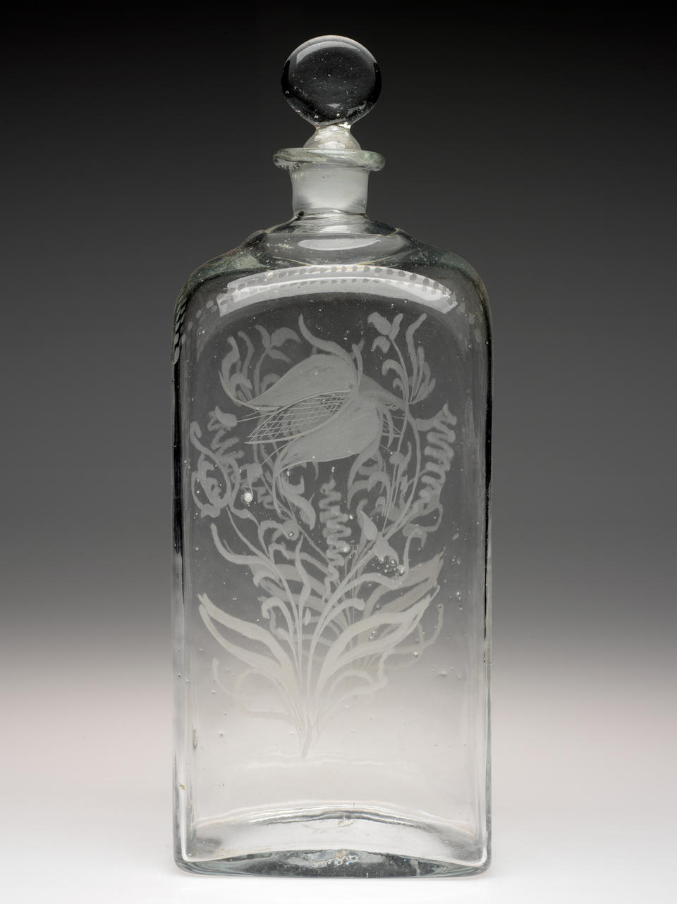 Tall rectangular clear bottle with rounded stopper. A floral design with leaves and swirling lines is etched on the front and back. 