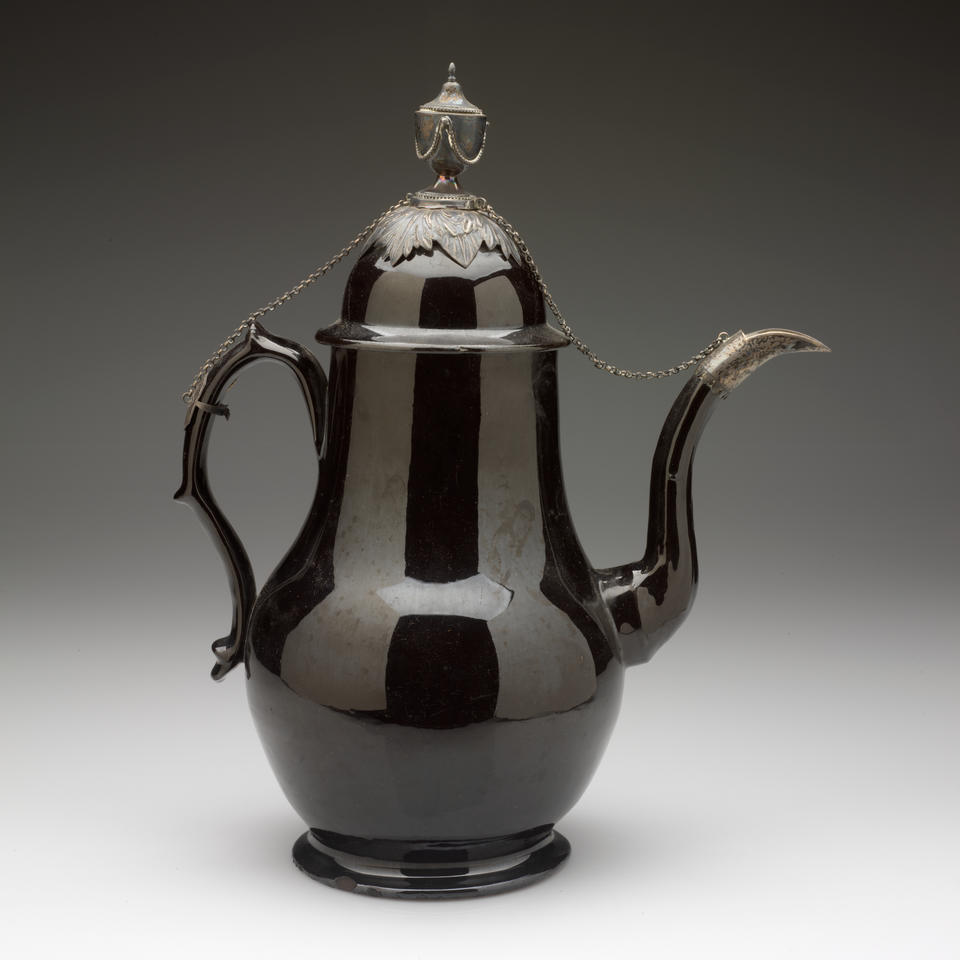 A glazed black teapot with a decorative handle, long curved spout, and a short foot, metal decorations on the spout, lid, finial, and handle with a chain connecting them. 